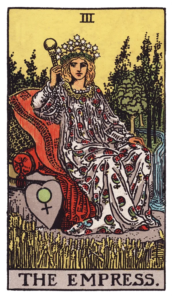 The Empress from the Rider-Waite-Smith deck: a pregnant-looking woman in a flowing gown patterned with pomegranates reclines on a cushioned outdoor throne set in a lush wheat field, a crown of twelve stars on her head, a heart-shaped shield bearing the astrological symbol for Venus resting beside her, and a stream of water flowing through trees in the background.