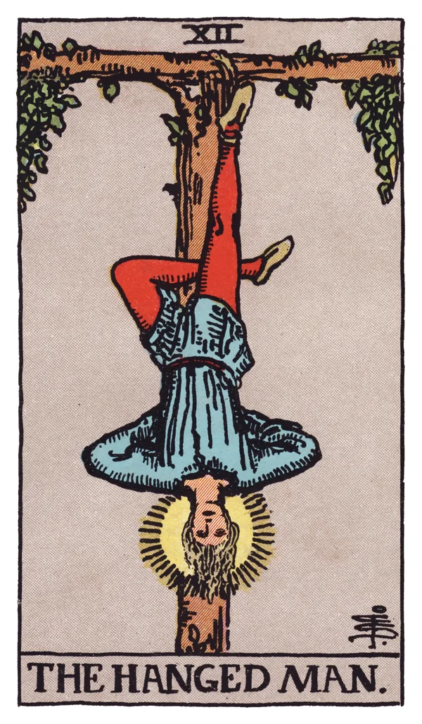 The Hanged Man from the Rider-Waite-Smith deck: a young man hangs upside down by one foot from a T-shaped wooden gibbet formed from a living tree, his other leg bent to form a numeral four, his hands folded calmly behind his back, a soft golden halo glowing around his head, his expression peaceful rather than distressed.