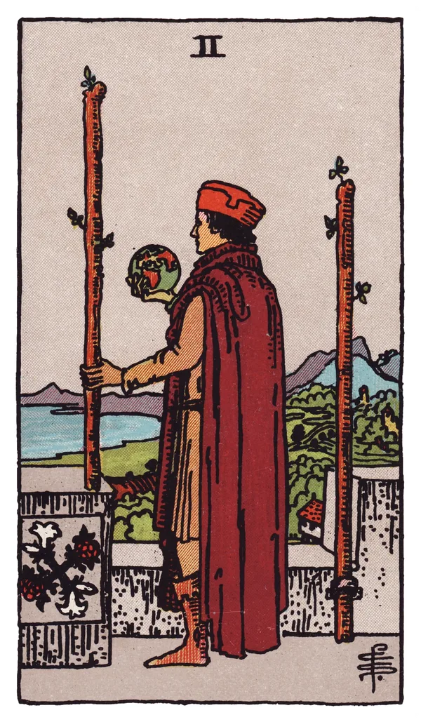 The Two of Wands from the Rider-Waite-Smith deck: a man in rich robes stands on a castle battlement overlooking sea and distant mountains, one wand fixed to the wall beside him and the other held loosely in his hand, a small globe resting in his other palm.